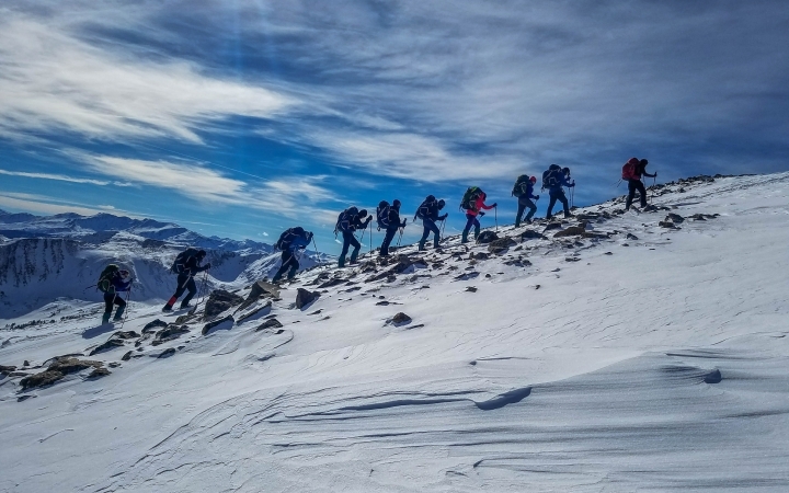 A group of gap year students hike along a snowy ridge. There are mountains in the background. 